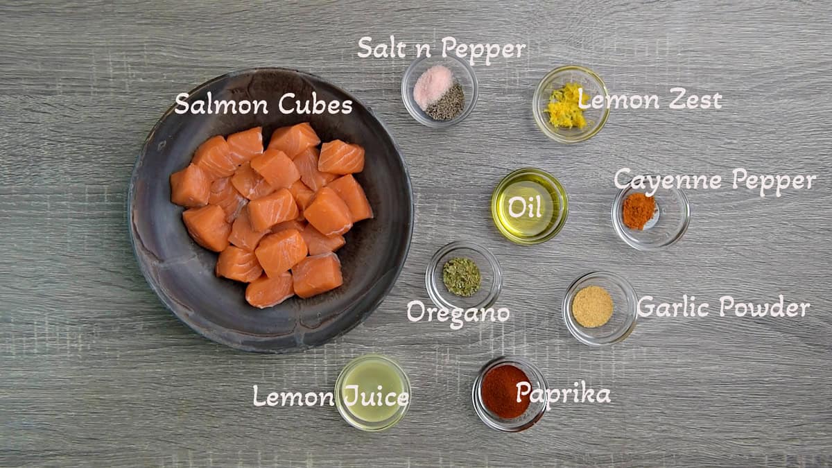 Recipe ingredients, each in individual bowls: Raw salmon cubes, salt and pepper, lemon zest, oil, cayenne pepper, garlic, paprika, oregano, lemon juice.