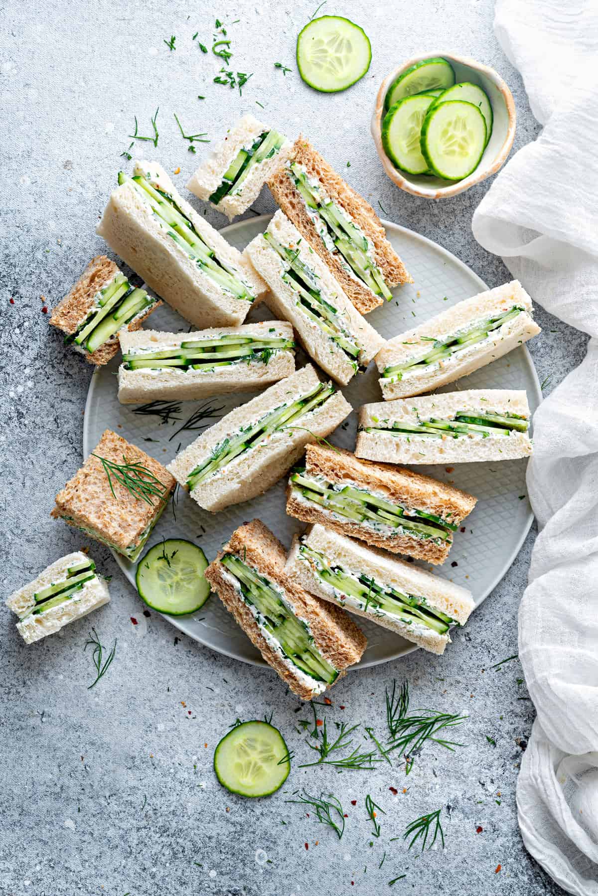 Top down view of a dozen cucumber cream cheese finger sandwiches laying all different directions on a large gray plate.