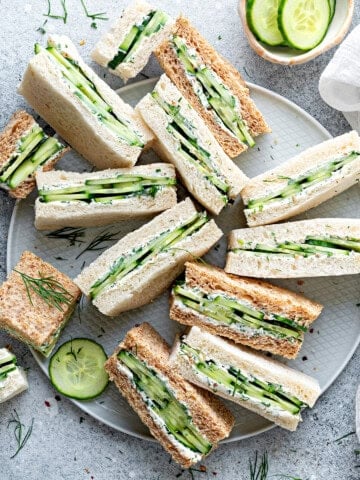 Top down view of a dozen cucumber and cream cheese finger sandwiches laying all different directions on a large gray plate.