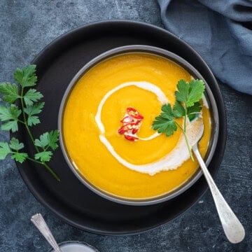 A bowl of pumpkin curry soup with herbs, red chili peppers for garnish, and coconut milk drizzled on top.