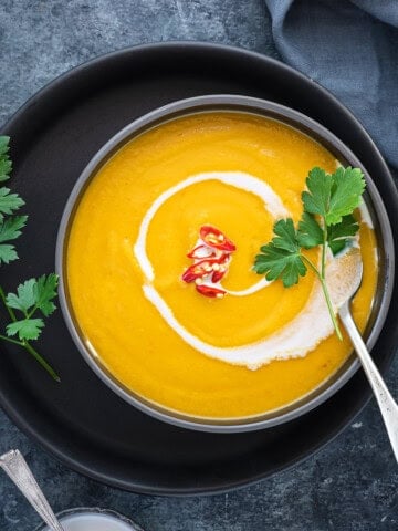 A bowl of pumpkin curry soup with herbs, red chili peppers for garnish, and coconut milk drizzled on top.