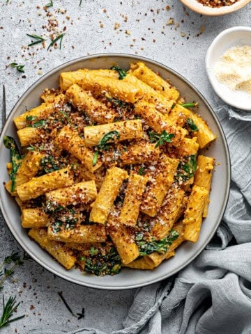 A bowl of creamy pumpkin pasta with spinach, parmesan, and red pepper flakes in bowls on the side.