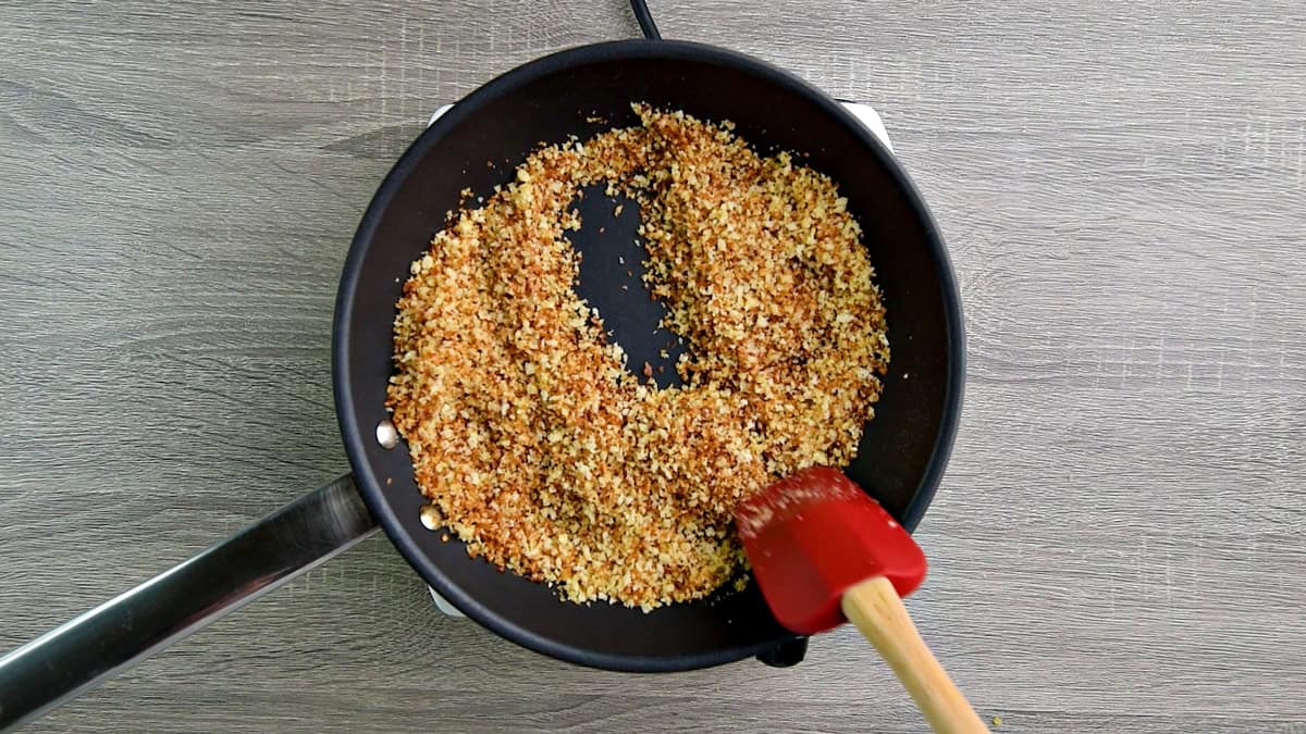 Toasted breadcrumbs being stirred in a skillet by a red spatula.