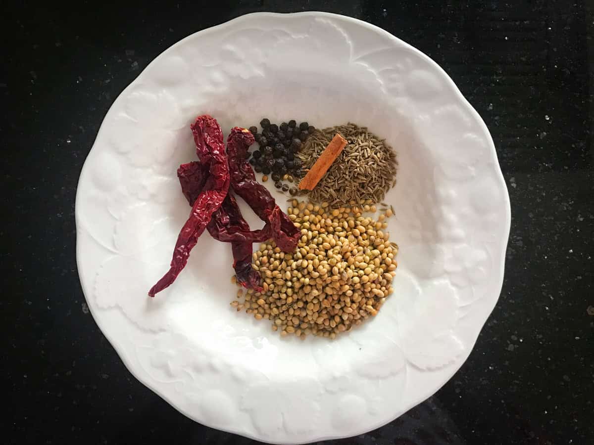 Coriander seeds, dry red chilies, cinnamon stick, black peppercorns and cumin seeds in a white ceramic plate.