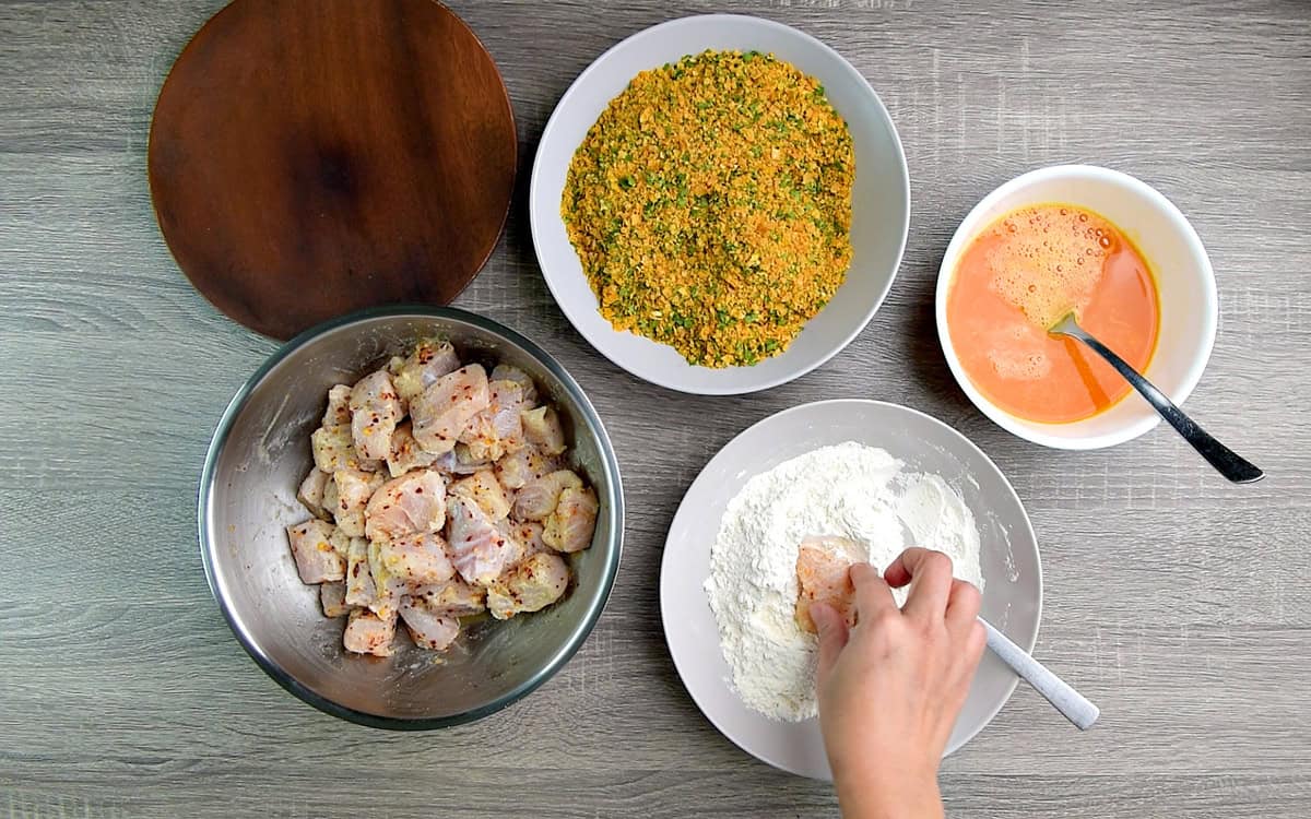 All the bowls of the step-by-step process shot with a hand coating the marinated fish in seasoned flour.